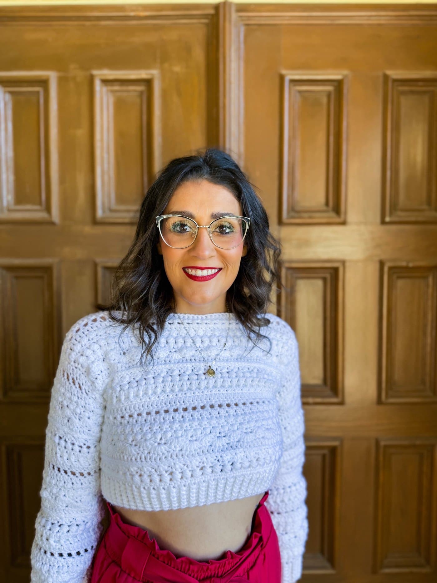 Chelsye Knox smiling in glasses and red lipstick, wearing a white crop sweater she crocheted, in front of wooden paneled doors
